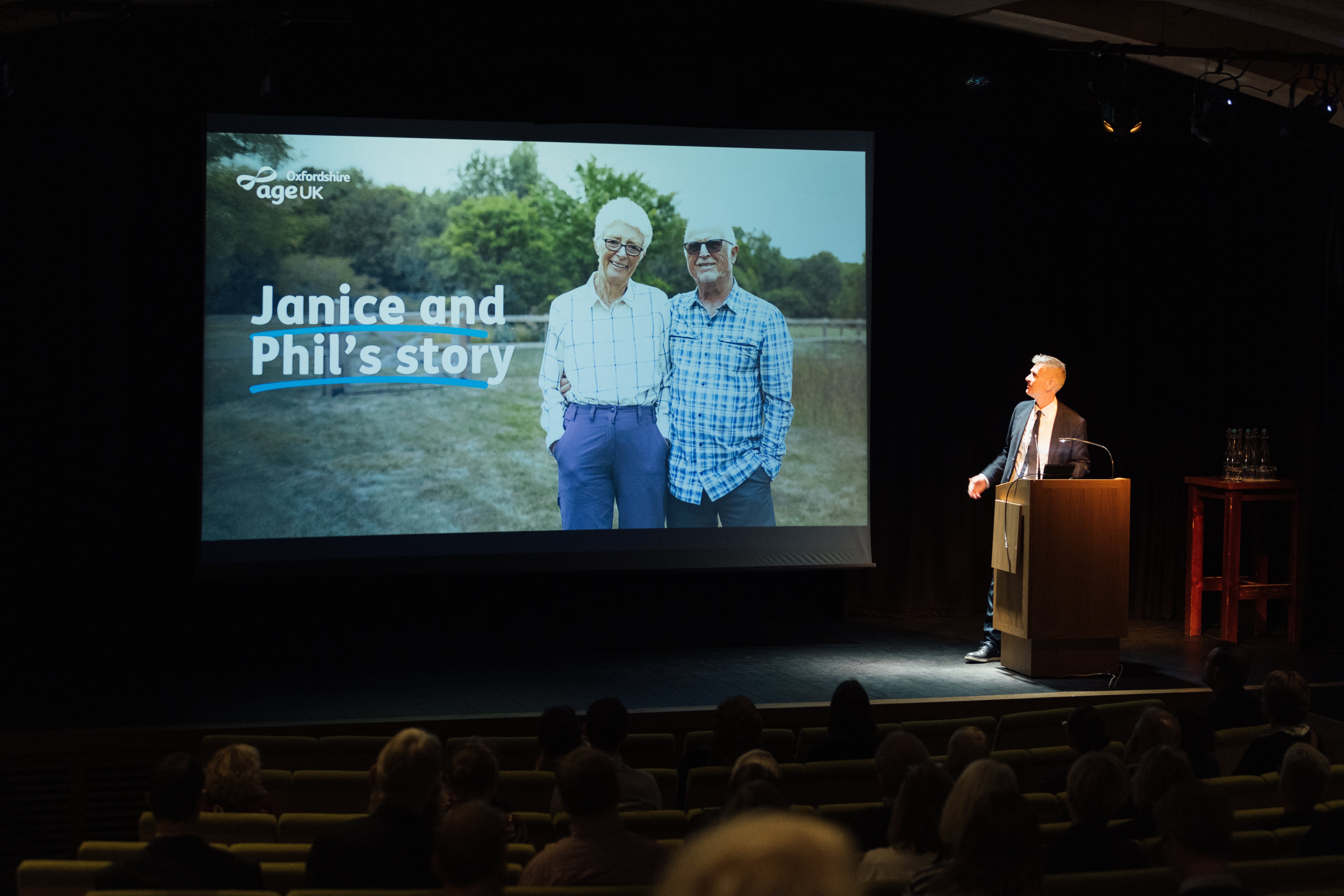 Photograph of Paul Ringer CEO pointing to the large screen which is showing the start of a film about a couple Janice and Phil.