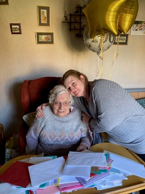 Connie sitting in an armchair at home, infront of many cards and envelopes. She is hugged by Sara, the community warden.