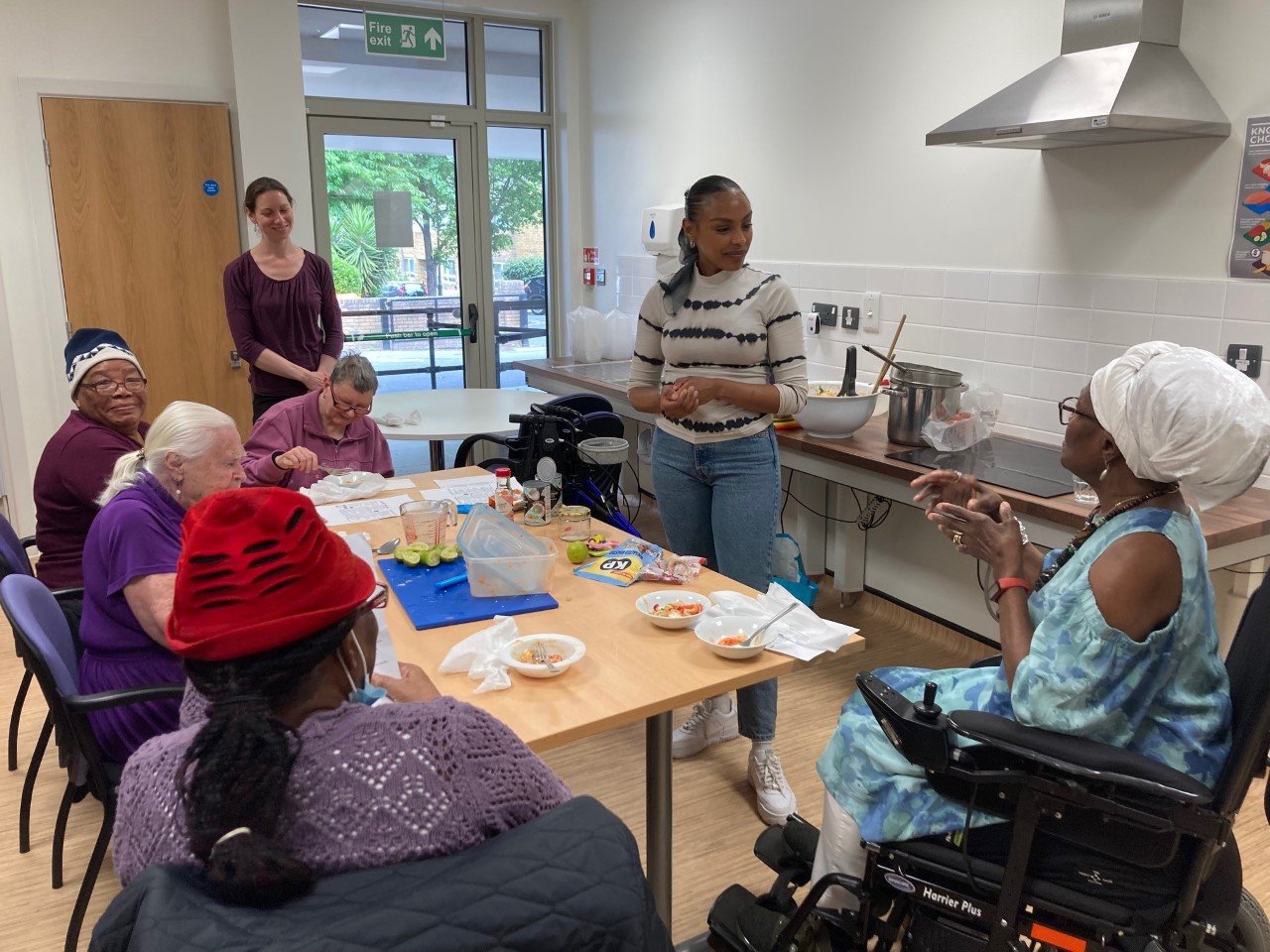 Picture of a group of service users and staff. They are sat round a table full of ingredients. They are listening to an instructor.