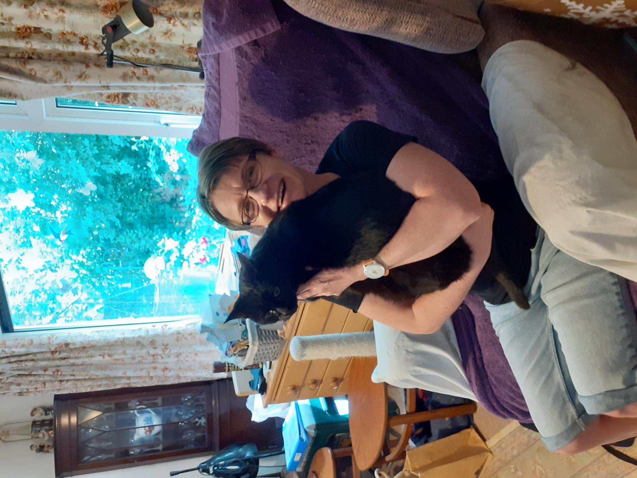 Anke, a lady sitting in an armchair holding a black cat on her lap, smiling at the camera.