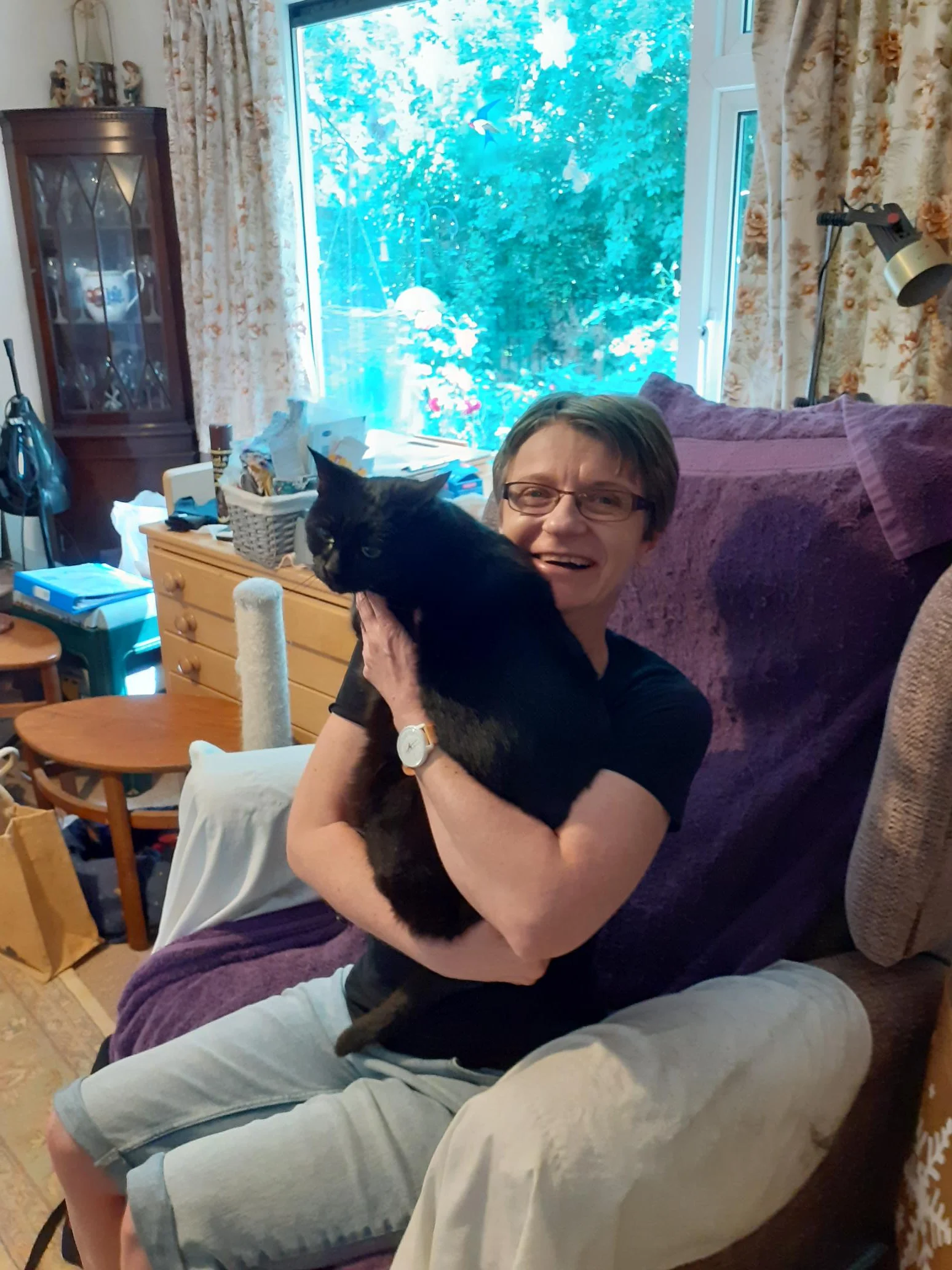 Anke, a lady sitting in an armchair holding a black cat on her lap, smiling at the camera.