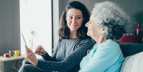 A younger white woman shows an older white woman something on a digital device.