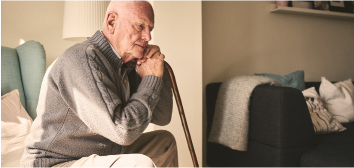 An older man leaning forward on a walking stick, whilst sitting in an armchair in his living room, staring sideways blankly.