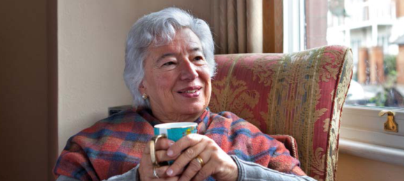older lady sitting in armchair with a warm blanket over shoulders and holding a mug of warm drink, smiling 