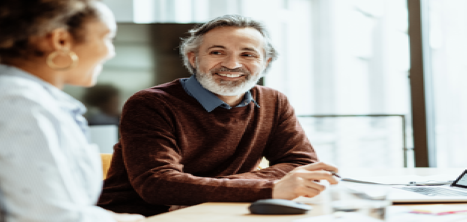 Older man in office at desk with laptop smiling and speaking to female colleague