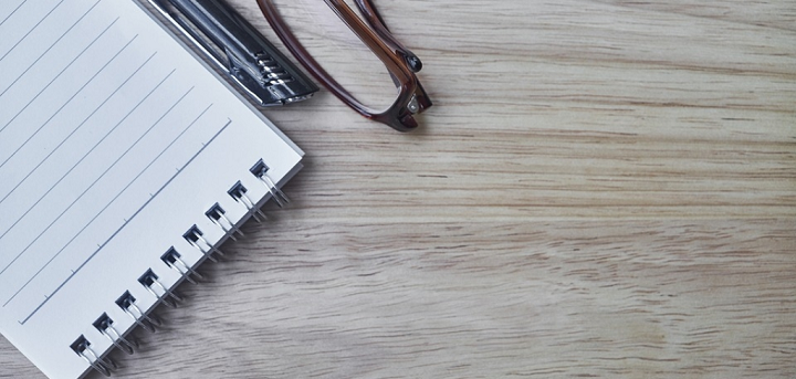 A pair of glasses and a notebook lying on a desk.