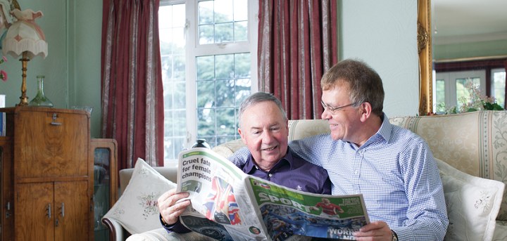 Two older men sat on the sofa reading the paper together. The man on the right has his arm around the man on the left.