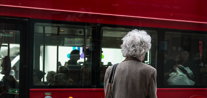 An older woman stood in front of a red London bus. She is facing away from the camera. Freedom Pass