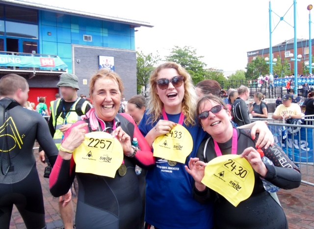 Three women in swimming suits laughing, displaying their swimming hats for the Crossacres Cruisers's Great Swim Manchester '15 event