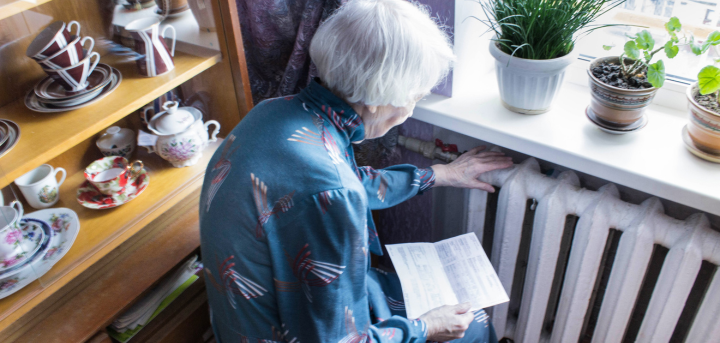 Older woman with her hand resting on a radiator, she has a utility bill in her other hand.