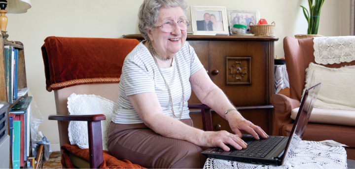 An older lady sits on a chair in her living room. She is using her laptop and smiling.