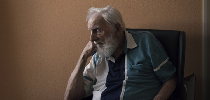 An older man sits in an armchair, looking pensive