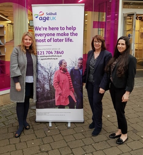 Three female presenting people in business attire standing by an Age UK Solihull sign that reads "We're here to help everyone make the most of later life."