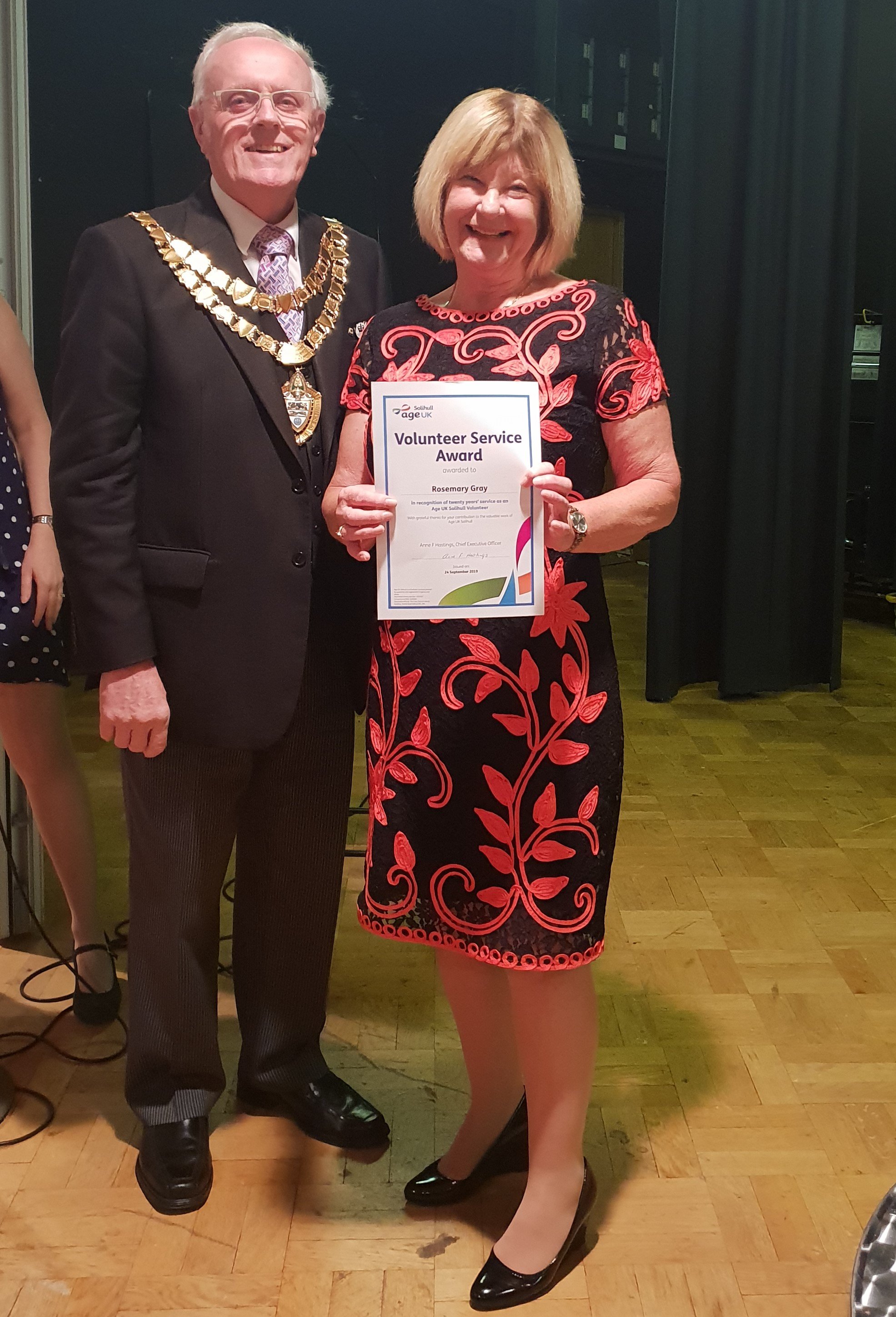 Mayor of Solihull stands with Rosemary Gray, who is wearing a black and red dress and holding a certificate for 20 years' service as an Age UK Solihull volunteer.