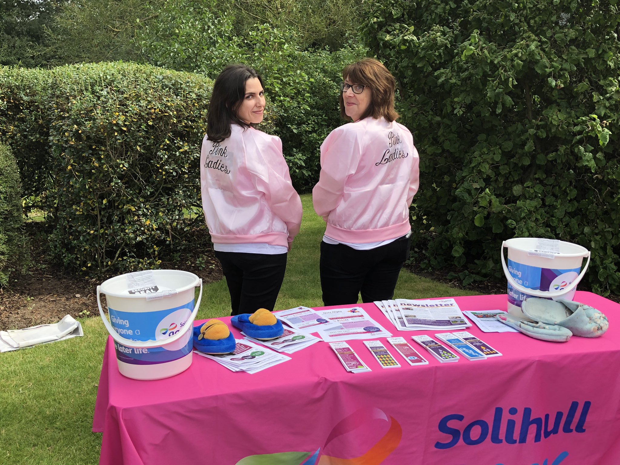 Two female presenting people wearing pink jackets, standing at a table covered in a pink table cloth displaying Age UK Solihull marketing materials