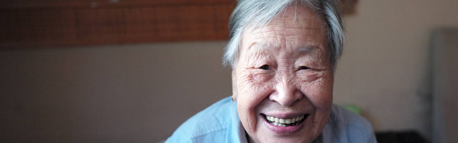 An older Asian man sits on his bed, smiling broadly
