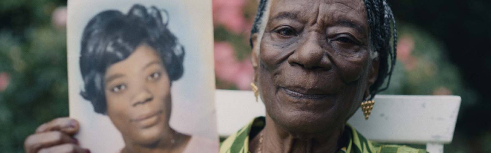 Linneth, an older Black woman, holds up a photo of herself from when she was young