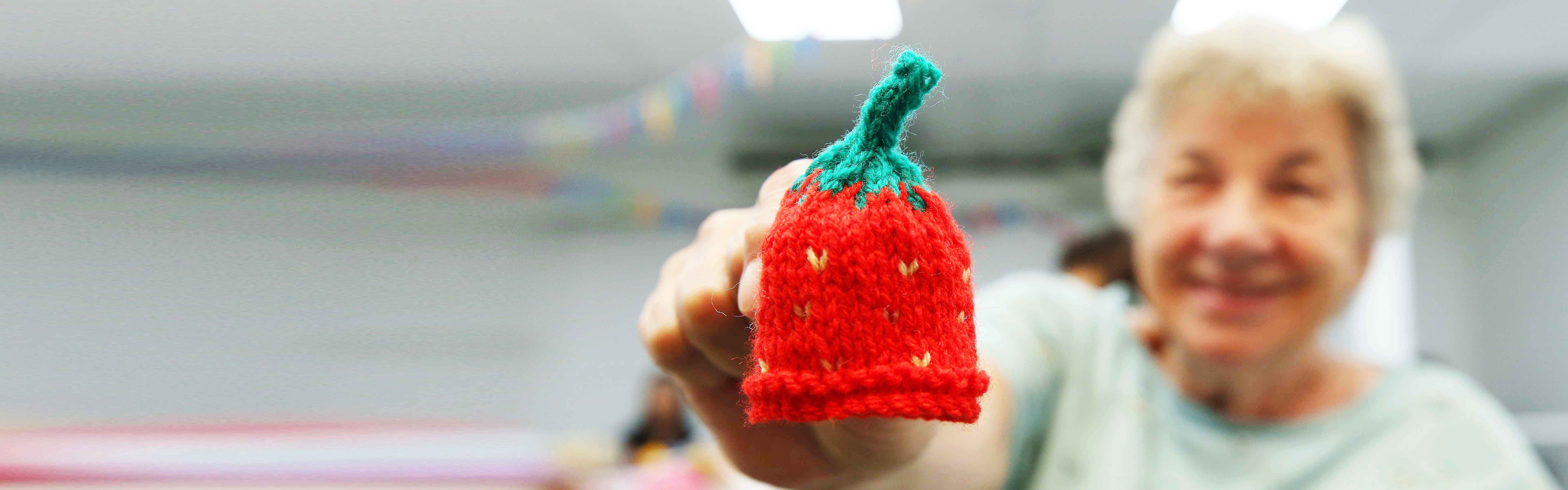 an older lady holds up a small knitted hat to the camera