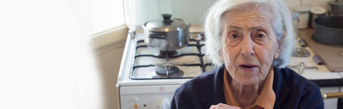An older woman sits in her kitchen, looking concerned