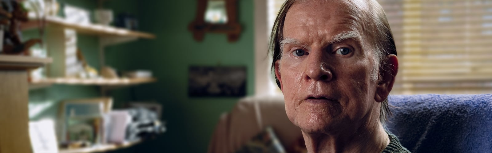 An older man sits in an armchair, looking sadly at the camera
