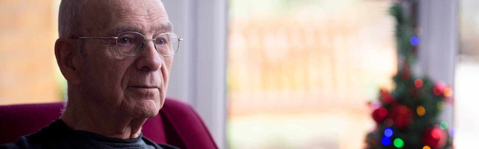 An older man, Roy, sits in his living room next to a small Christmas tree, looking wistful