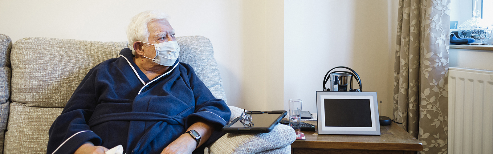 An older man, sat on a sofa and wearing a protective face mask