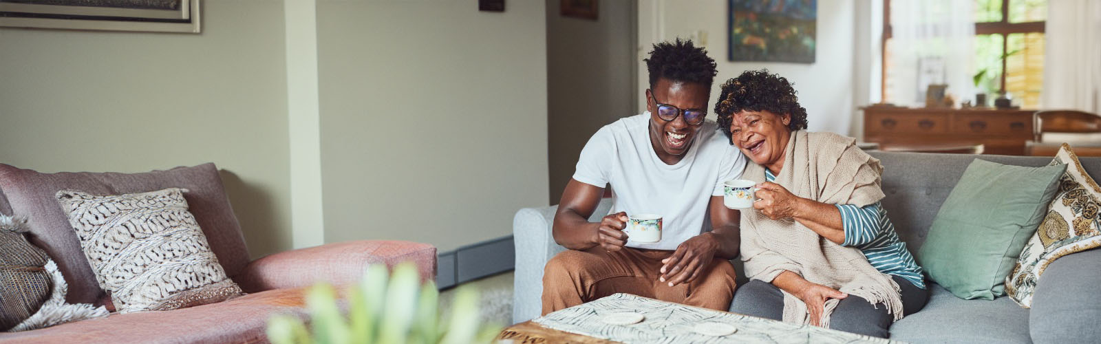 A younger man and older woman sit on the sofa with cups of tea laughing