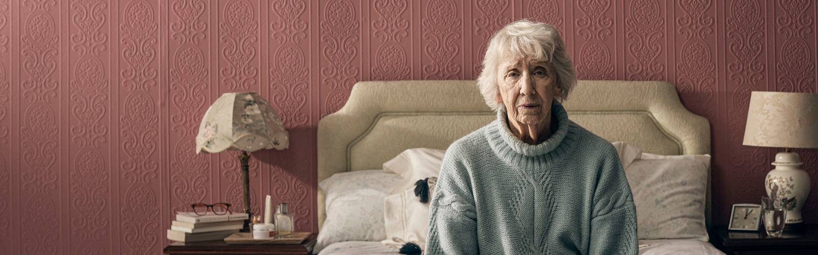 An older woman sits alone on her bed, looking worried