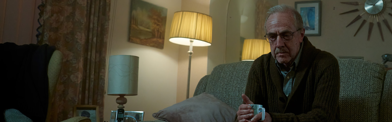 An older man with glasses sits alone on a sofa in the dark, looking sadly off camera