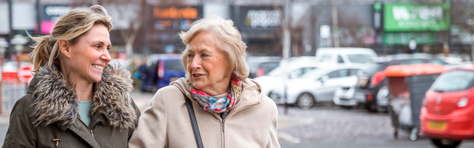 A younger woman and her older female relative walk arm in arm through a supermarket car park