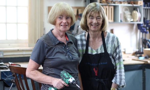 Two older women in a carpentry workshop smile at the camera