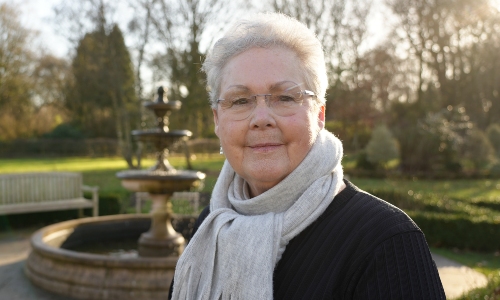 An older lady with short hair and glasses stands in a park, looking to camera