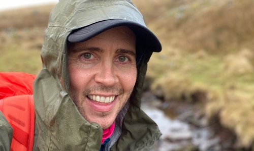 A man dressed in an anorak and outdoor gear smiles while hiking in the rain