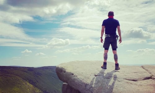 A man stands on a cliff edge, looking over the mountains below