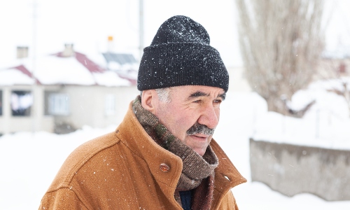 An older man with a moustache and a beanie hat, looking cold against a snowy background