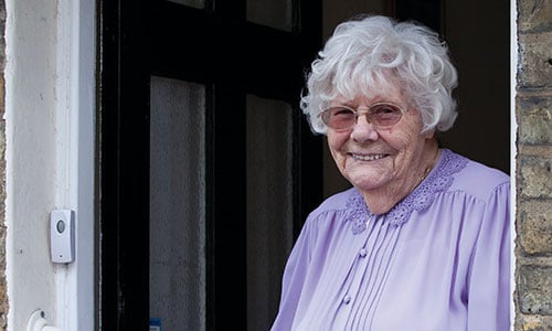 An older woman looking out from the front door of her house