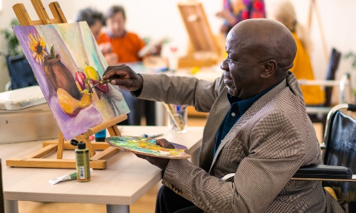 An older Black man sits painting on an easel
