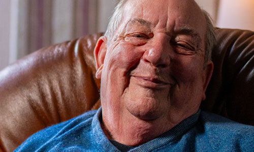 An older man in a blue jumper, sat in a chair, smiling