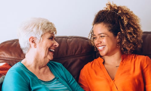 An older woman and younger woman sit next to each other on a sofa, laughing