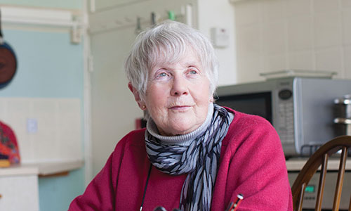 An older woman wearing a red jumper and scarf, sat in her kitchen