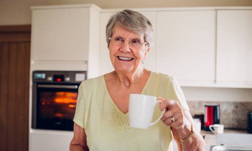 An older woman wearing a red jumper and scarf, sat in her kitchen