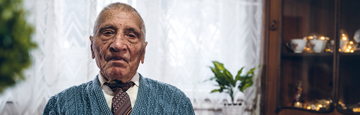 An older man looks directly at the camera in his house.