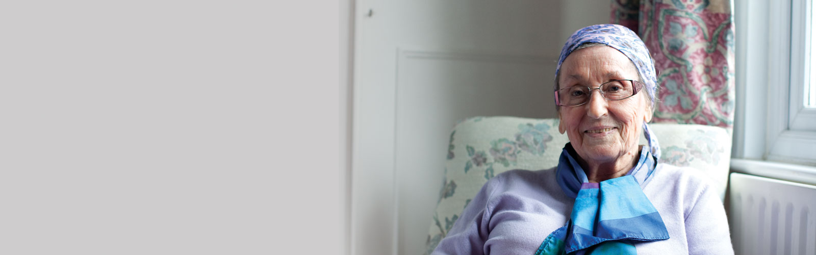 An older lady sits at home, wearing a scarf and glasses