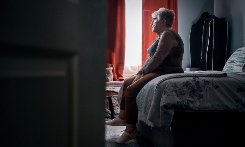An older woman sits on a bed in the dark, looking lonely