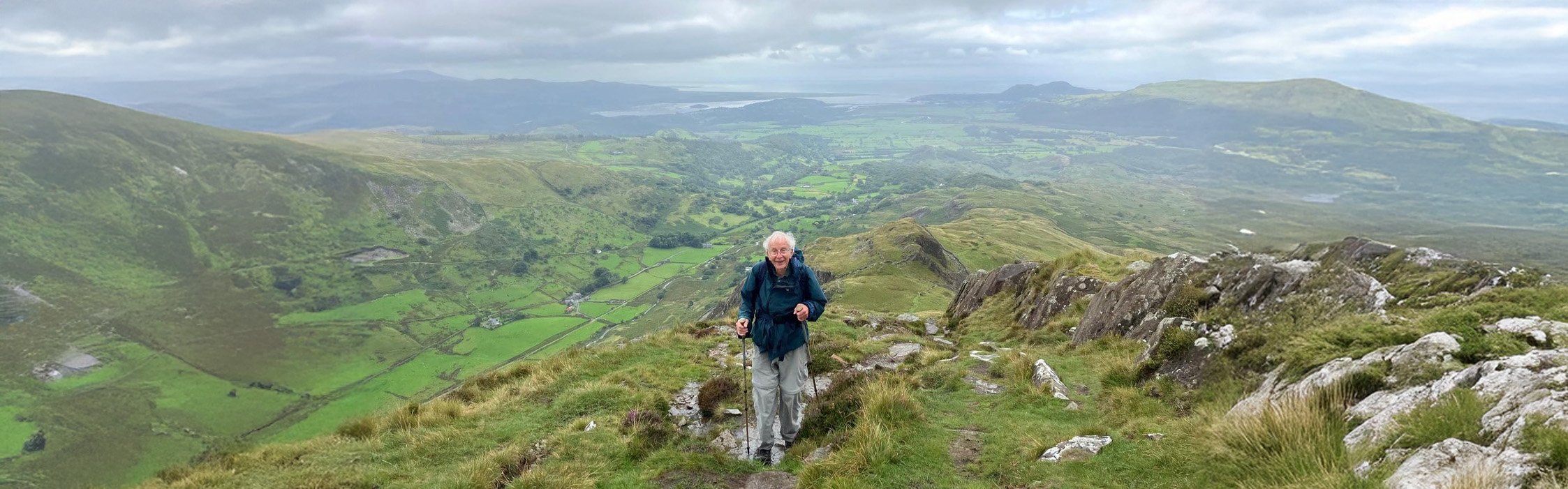 An older man, in the centre of the image, walking against the green hills of the Peak District