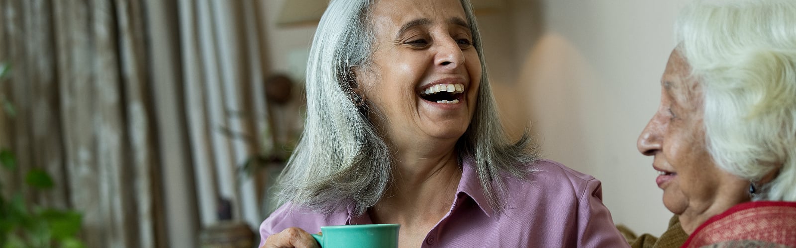 Two older women of South Asian heritage laughing over a cup of tea