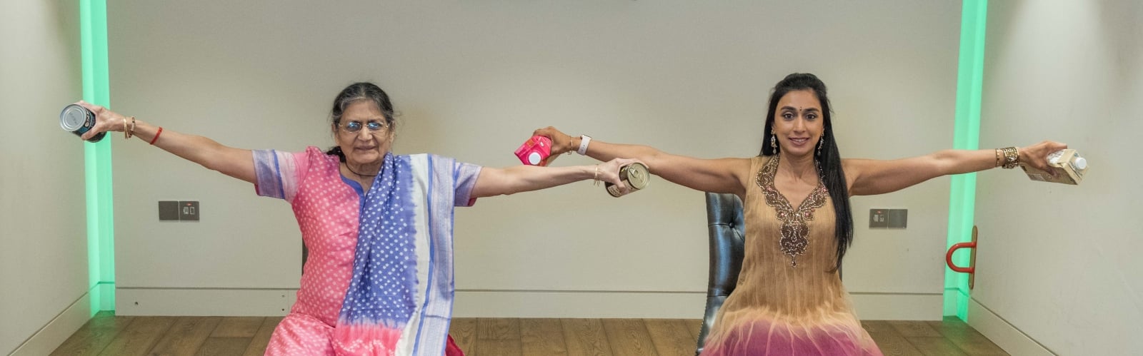 An older South Asian woman pictured with a younger South Asian woman, both sat doing arm exercises