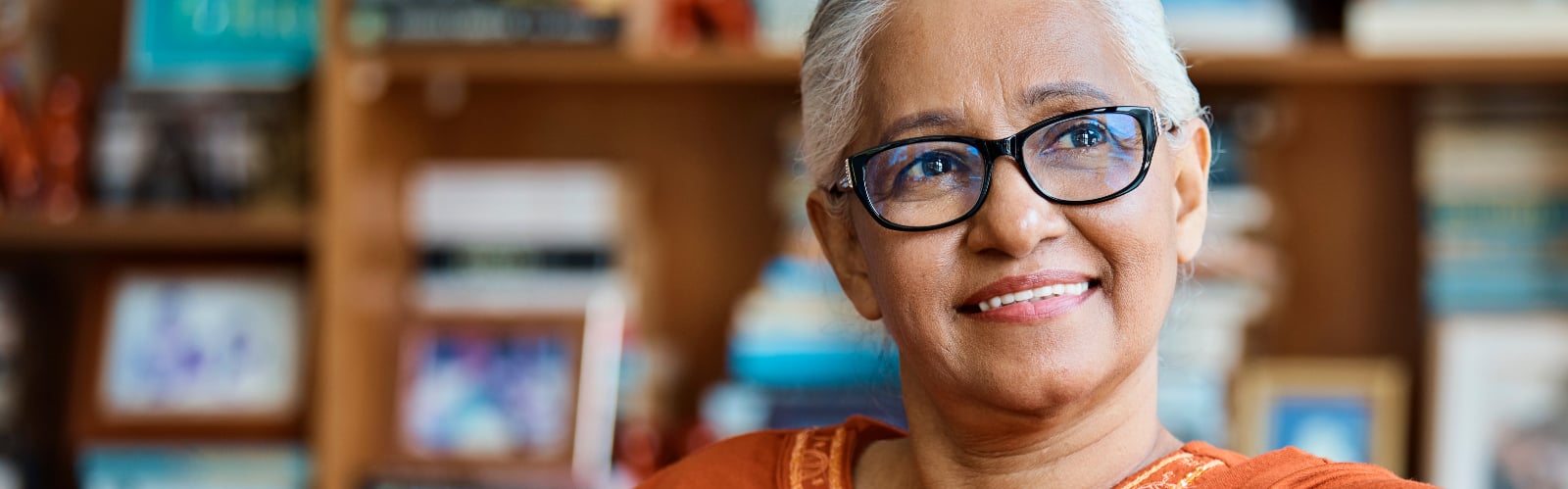 A middle-aged woman with short brown hair sits holding a cup of tea and looking out of her window, smiling slightly