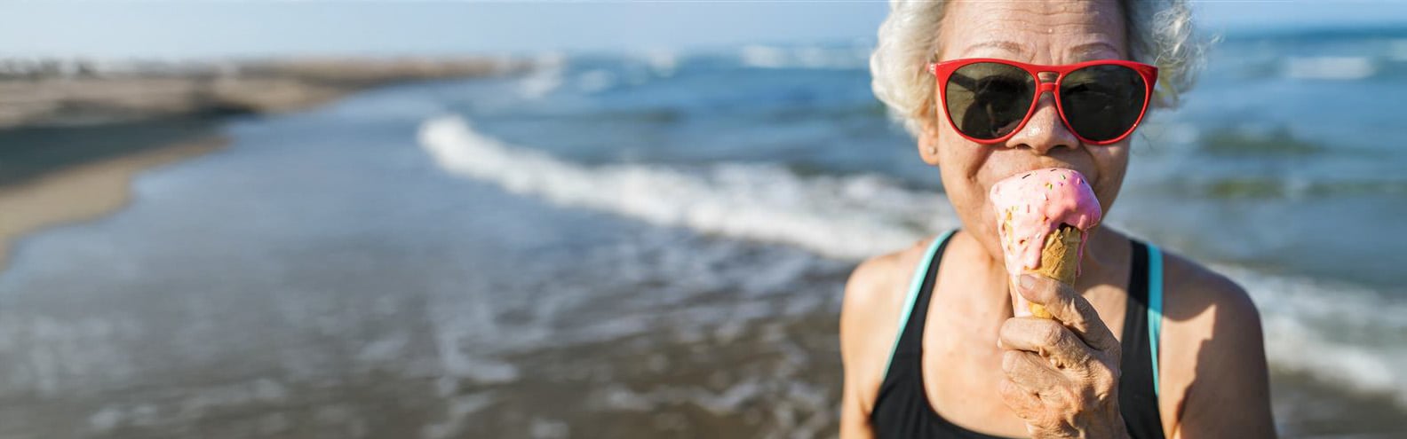 An older lady with sunglasses eats an ice cream by the sea