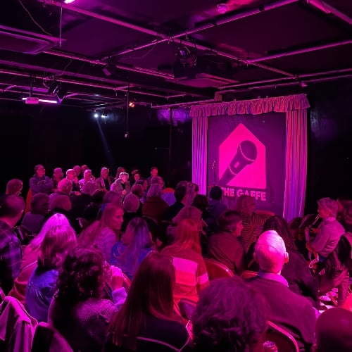 An audience in a comedy club, bathed in a pink light as they await the comedians